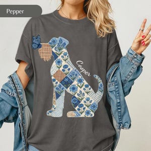 May include: A charcoal grey t-shirt featuring a graphic of a dog silhouette filled with a patchwork pattern of blue flowers, brown plaid, and light blue gingham. A blue butterfly rests on the dog's nose, and the name Cooper is written in white.