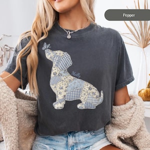 May include: Dark gray t-shirt featuring a patchwork dog design with blue floral and gingham patterns. A butterfly rests on the dog's nose. The name "Molly" is written on the shirt. The word "Pepper" is on a gray bar.