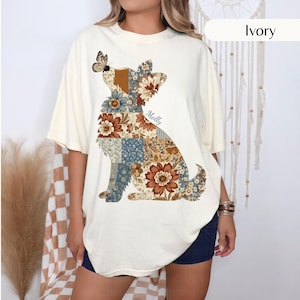 May include: An ivory short-sleeve t-shirt featuring a large graphic of a dog silhouette filled with a patchwork pattern of brown, blue, and cream floral designs. A small butterfly rests on the dog's head, and the name Molly is printed in script.