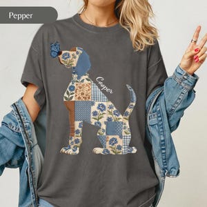 May include: A charcoal grey t-shirt featuring a graphic of a dog composed of various blue and tan floral and checkered quilt-style patches. A blue butterfly rests on the dog's nose, and the name Cooper is printed in white script above the tail.