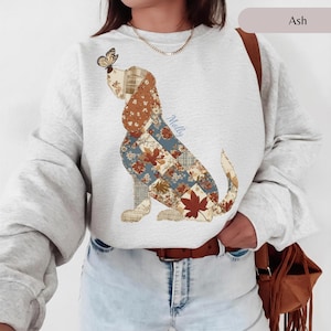 May include: An ash gray crewneck sweatshirt featuring a patchwork-style dog illustration with floral and plaid patterns in shades of brown, blue, and cream. A butterfly rests on the dog's nose, and the name Molly is printed in blue script.