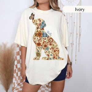 Puede incluir: Una camiseta color marfil con un gráfico grande de un perro sentado compuesto por varios patrones florales y de cuadros en tonos naranja, azul y marrón, con una mariposa en la nariz y el nombre Molly impreso en letra cursiva azul.