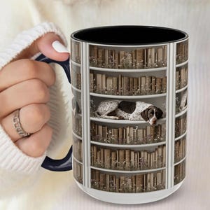May include: White ceramic mug with a wraparound design of a dog resting on a bookshelf filled with vintage books. The mug has a black interior and a navy blue handle. The design includes floral accents.