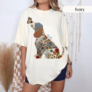 May include: An ivory short-sleeve t-shirt featuring a graphic of a dog composed of various floral and plaid fabric-style patterns. The dog has a brown ear, a butterfly on its nose, and the name Molly written in blue script near its back.