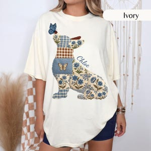 以下が含まれることがあります： アイボリー色のTシャツ。青い花柄、茶色のギンガムチェック、青い格子柄などのパッチワーク模様で構成された犬のグラフィックが描かれています。犬の頭には青い蝶がとまっており、青い筆記体でChloeという名前がプリントされています。