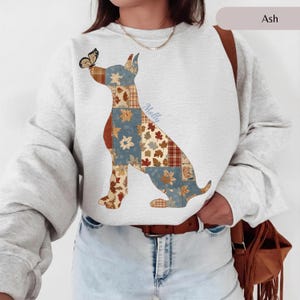Puede incluir: Una sudadera gris ceniza de cuello redondo con un gráfico grande de un perro sentado. El perro está compuesto por varios cuadrados de patchwork con patrones de hojas de otoño en azul, naranja y marrón. Una mariposa descansa sobre su nariz.