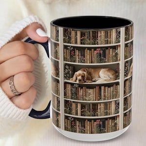 May include: A white ceramic mug with a black interior and a blue handle. The mug features a design of a bookshelf filled with books and flowers, with a sleeping dog in the center. The mug is held by a person.