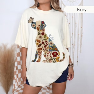 May include: An ivory short-sleeve t-shirt featuring a graphic of a sitting dog composed of various floral and leaf patterns in shades of red, blue, yellow, and green. A butterfly rests on the dog's nose, and the name Molly is printed in script.
