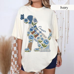 May include: An ivory t-shirt featuring a graphic of a dog silhouette filled with a blue floral and plaid patchwork pattern. A small blue butterfly rests on the dog's nose, and the name Cooper is printed in blue script next to the dog's back.