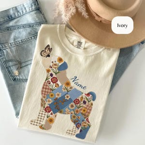 May include: An ivory t-shirt featuring a graphic of a dog silhouette filled with a patchwork pattern of blue, brown, and floral squares. A butterfly rests on the dog's head. The text 'Name' is printed in blue script. A tan hat and blue jeans are visible.