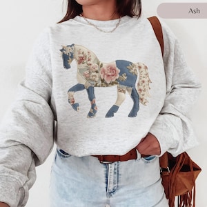 May include: Light grey sweatshirt with a floral horse motif. The horse is made from patchwork fabric in blue, cream, and pink, with a large rose in the center. The sweatshirt has long sleeves and a crew neck.