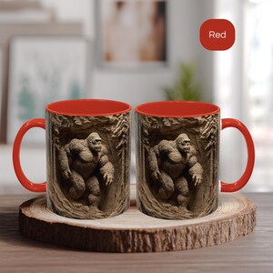 May include: Two red-handled coffee mugs with a brown exterior featuring a detailed relief of a Bigfoot figure in a cave setting. The interior of the mugs is red. The mugs are displayed on a wooden coaster.