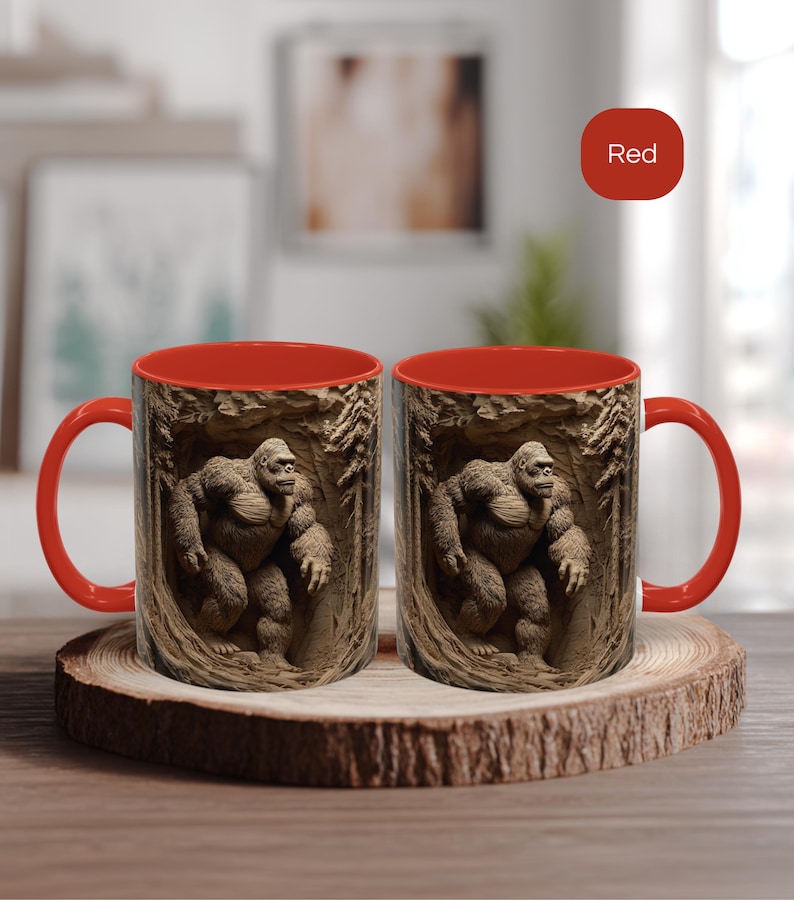 May include: Two red-handled coffee mugs with a brown exterior featuring a detailed relief of a Bigfoot figure in a cave setting. The interior of the mugs is red. The mugs are displayed on a wooden coaster.