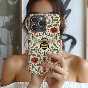 May include: A phone case featuring a bee design. The case has a cream background with red flowers and green leaves. The bee is centered with black and yellow stripes and detailed wings. The case is designed for a smartphone.
