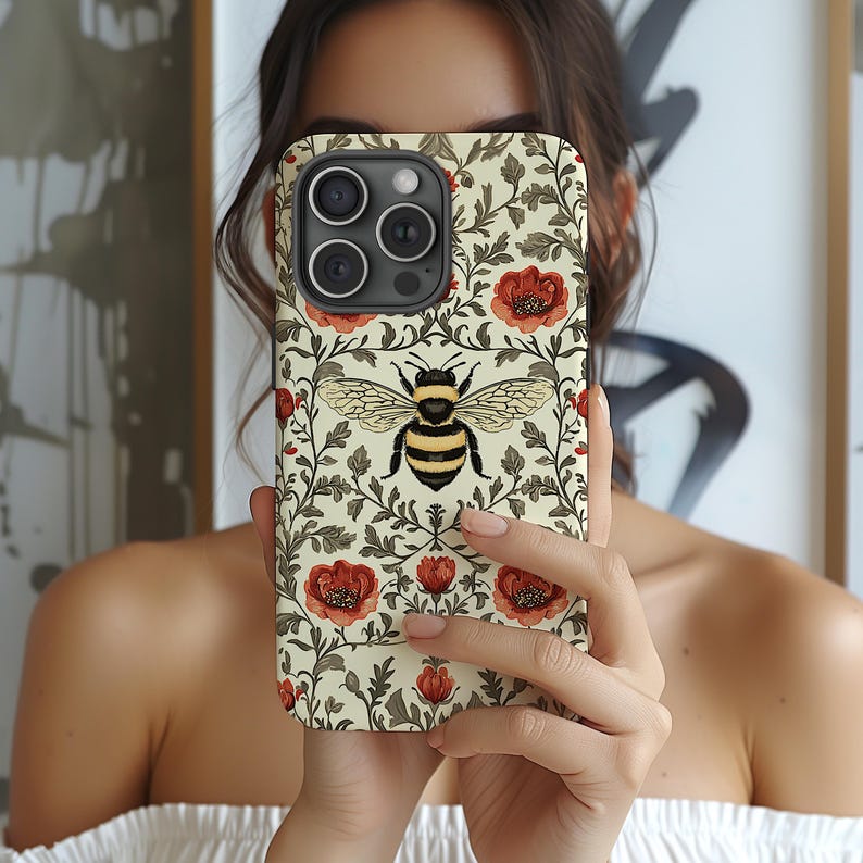 May include: A phone case featuring a bee design. The case has a cream background with red flowers and green leaves. The bee is centered with black and yellow stripes and detailed wings. The case is designed for a smartphone.