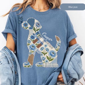 May include: A blue jean colored t-shirt featuring a graphic of a dog silhouette filled with a blue floral and plaid patchwork pattern. A blue butterfly rests on the dog's nose, and the name Cooper is written in white script next to the dog.