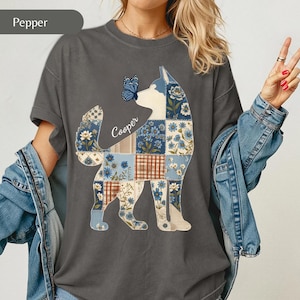 May include: A charcoal grey t-shirt featuring a graphic of a dog silhouette filled with a blue, white, and brown floral patchwork pattern. A blue butterfly rests on the dog's nose, and the name Cooper is printed in white script next to the dog.
