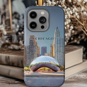 May include: A phone case featuring a Chicago cityscape with the iconic Cloud Gate sculpture. The design includes the word "CHICAGO" in a bold font, set against a backdrop of skyscrapers and a blue sky.