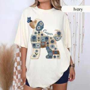 May include: An ivory short-sleeve t-shirt featuring a graphic of a dog composed of blue and white floral patchwork squares. The dog design includes a blue butterfly on its head and the name Cooper written in blue script text.