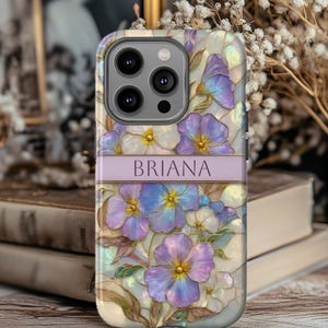 May include: A phone case featuring a floral design with purple, blue, and white flowers. The case has a pearlescent background and a lavender band with the name "BRIANA" printed on it. The case has a camera cutout.