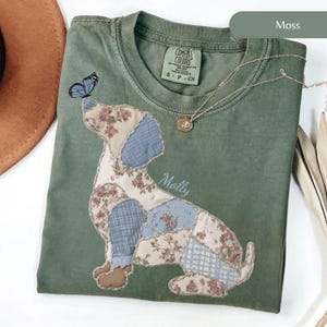 May include: Moss green t-shirt featuring a patchwork dachshund design with floral and blue patterned fabric. A blue butterfly rests on the dog's nose. The name "Molly" is written on the shirt. A gold necklace with a pendant is also visible.