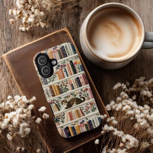 May include: A phone case with a bookshelf design, featuring a cat resting on a shelf with colourful books and flowers. The case is on a brown leather-bound book, beside a coffee cup and dried flowers.