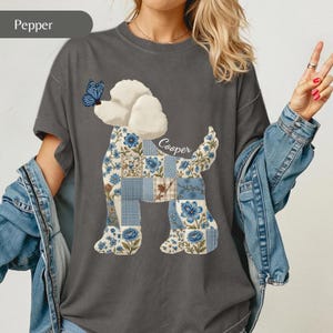 May include: A charcoal gray t-shirt featuring a graphic of a poodle dog with a patchwork body design in shades of blue and white floral and plaid patterns. A blue butterfly rests on the dog's nose, and the name Cooper is written in script.