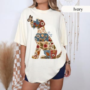May include: An ivory short-sleeve t-shirt featuring a graphic of a dog silhouette composed of various floral and leaf-patterned patches in shades of blue, red, and orange. A butterfly rests on the dog's nose, and the name Molly is printed nearby.