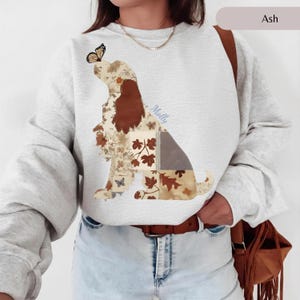 May include: An ash gray crewneck sweatshirt featuring a graphic of a dog silhouette composed of various patchwork patterns in shades of brown, cream, and tan. The design includes autumn leaf motifs, a butterfly on the dog's nose, and the name Molly.