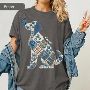 May include: A charcoal grey t-shirt featuring a graphic of a dog silhouette filled with a blue and white floral and plaid patchwork pattern. A blue butterfly rests on the dog's nose, and the name Cooper is printed in white script next to the dog.