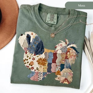 May include: A moss green t-shirt with a patchwork dog design. The dog is made of various fabric patterns, including floral and striped designs. The shirt has a crew neck and a small tag that says "Comfort Colors". A silver necklace with a pendant is also visible.