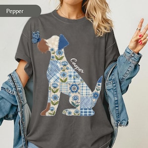 May include: A charcoal grey t-shirt featuring a graphic of a dog composed of blue and white floral and plaid patchwork patterns. The dog has a brown ear, a blue ear, and a blue butterfly on its nose. The name Cooper is printed in white script.