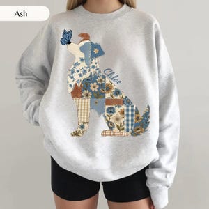 May include: An ash gray crewneck sweatshirt featuring a large graphic of a dog composed of various blue, tan, and white floral and plaid patchwork patterns. A blue butterfly sits on the dog's nose, and the name Chloe is printed in blue script.
