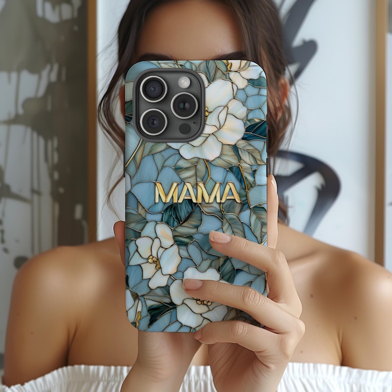 May include: A phone case with a floral design in shades of blue, white, and gold. The case features a mosaic-like pattern with flowers and leaves. The word 'MAMA' is displayed in gold letters across the center of the case. The phone case has three camera lenses.