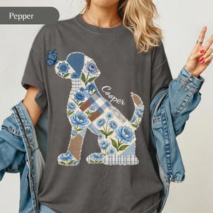 Puede incluir: Una camiseta gris carbón con un gráfico de la silueta de un perro relleno de un patrón de patchwork de flores azules, cuadros y secciones marrones sólidas. Una mariposa azul descansa sobre la nariz del perro y el nombre Cooper está escrito en blanco.