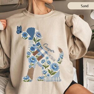 May include: A sand-colored crewneck sweatshirt featuring a large graphic of a dog silhouette composed of blue floral and plaid patchwork patterns. A blue butterfly rests on the dog's nose, and the name Cooper is printed in blue script.
