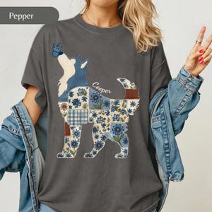 May include: A charcoal grey t-shirt featuring a graphic of a dog in a patchwork style with blue floral patterns, a solid blue head, and a brown tail. A blue butterfly rests on the dog's nose, and the name Cooper is written in white script.