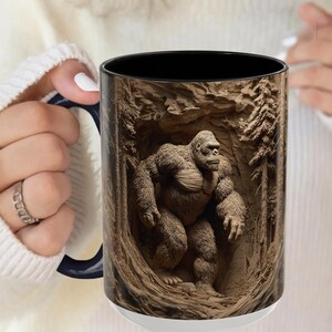 May include: White ceramic mug with a black interior and a detailed illustration of a gorilla in a forest setting. The gorilla is brown and the forest is carved into the mug's surface.