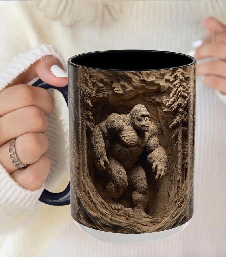 May include: White ceramic mug with a black interior and a detailed illustration of a gorilla in a forest setting. The gorilla is brown and the forest is carved into the mug's surface.
