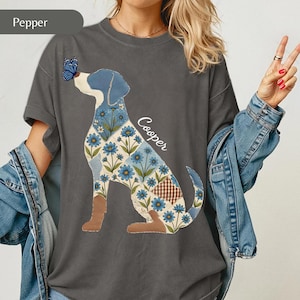 Puede incluir: Una camiseta gris carbón con el gráfico de un perro con un patrón de parches de flores azules y cuadros marrones. Una mariposa azul descansa sobre la nariz del perro y el nombre Cooper está impreso en letra blanca junto al perro.