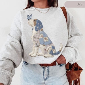 Puede incluir: Sudadera gris ceniza con un diseño de perro patchwork y una mariposa azul. El diseño del perro incorpora parches florales y a cuadros. La sudadera tiene cuello redondo y mangas largas.