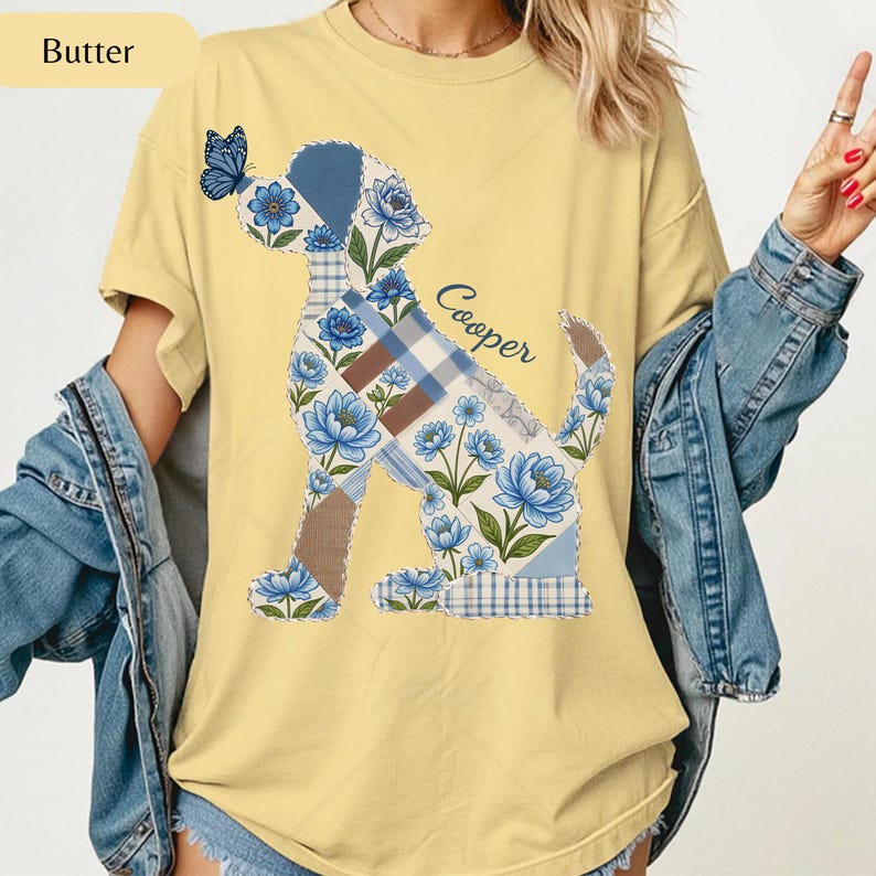 May include: A butter yellow t-shirt featuring a graphic of a dog silhouette filled with a patchwork pattern of blue flowers, plaid, and solid brown sections. A blue butterfly rests on the dog's nose, and the name Cooper is printed in blue script.