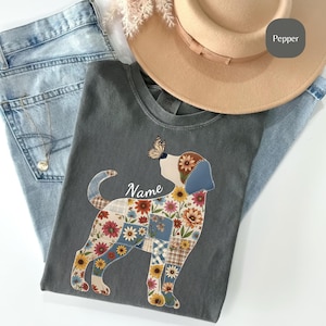 Puede incluir: Una camiseta gris carbón con la silueta de un perro rellena de un patrón de patchwork de girasoles, margaritas y tela de cuadros. Una mariposa descansa sobre la nariz del perro y la palabra Name está impresa en blanco sobre el lomo.