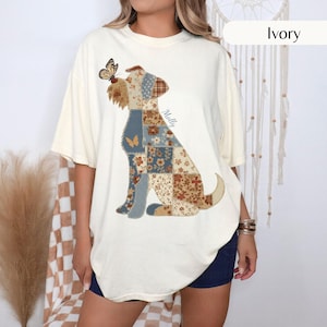 May include: An ivory short-sleeve t-shirt featuring a graphic of a dog composed of various brown, blue, and cream floral and geometric patchwork patterns. A butterfly rests on the dog's nose, and the name Molly is printed in script.