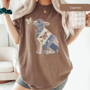 以下が含まれることがあります： エスプレッソ色のTシャツ。パッチワークの子羊のデザインと蝶が特徴です。子羊は青、白、花柄の生地で作られています。2匹目の蝶が子羊の頭の近くにいます。このシャツは快適でカジュアルなスタイルです。