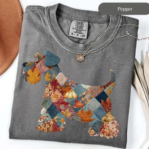 Peut inclure: Un t-shirt gris Comfort Colors avec un motif de chien en patchwork avec des motifs de feuilles d'automne. Le t-shirt a un col rond et des manches courtes. Un collier argenté avec un pendentif est drapé sur le t-shirt.