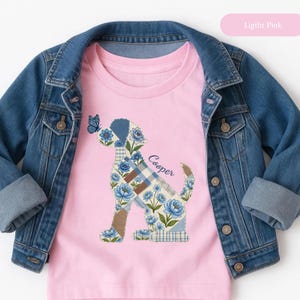 May include: A light pink t-shirt featuring a graphic of a dog silhouette filled with a blue floral and plaid patchwork pattern, with a small blue butterfly on the dog's nose and the name Cooper written in cursive script, layered under a denim jacket.