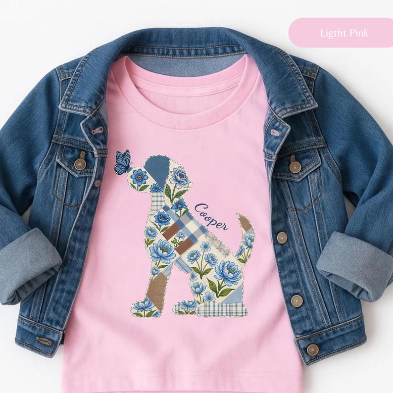 May include: A light pink t-shirt featuring a graphic of a dog silhouette filled with a blue floral and plaid patchwork pattern, with a small blue butterfly on the dog's nose and the name Cooper written in cursive script, layered under a denim jacket.
