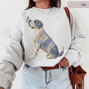 May include: Ash-colored sweatshirt featuring a whimsical illustration of a dog with patchwork quilt-style patterns in blue, pink, and cream. A butterfly rests on the dog's nose. The sweatshirt has long sleeves and a crew neck.