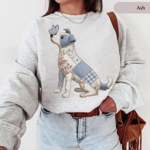 Peut inclure: Sweat-shirt gris cendré avec un motif de chien patchwork. Le chien présente des motifs floraux et à carreaux bleus, avec un papillon sur le nez. Le sweat-shirt a des manches longues et un col rond.